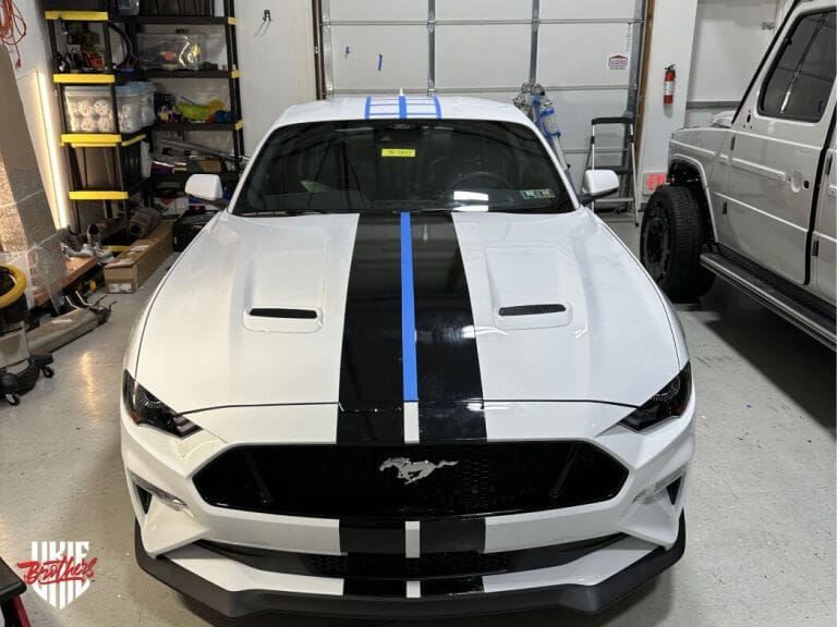 White Ford Mustang with a custom black racing stripe being applied by Ukie Brothers in a professional garage, showcasing a high-precision vinyl wrap installation