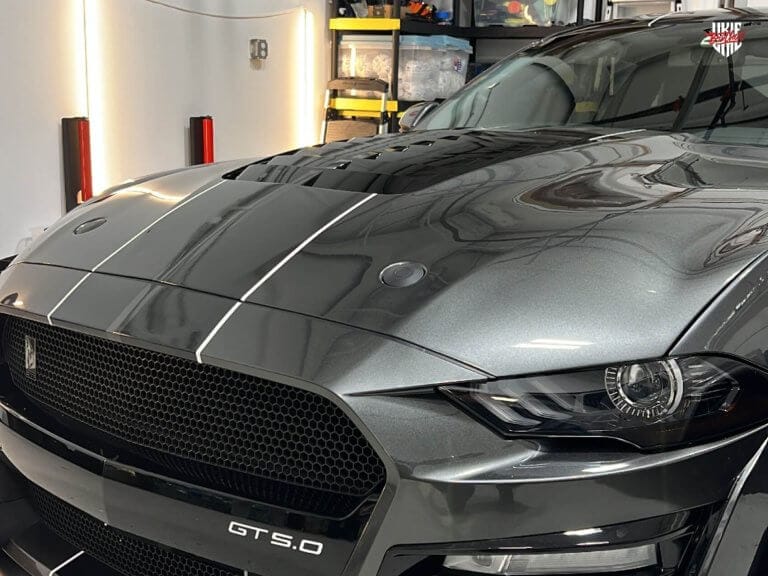 Grey Ford Mustang GT 5.0 with custom black and silver racing stripes vinyl wrap by Ukie Brothers, showcasing a sleek and aggressive front-end design in a professional garage.