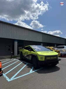 Professional Tesla Wrap Services – Model 3, Y, S & Cybertruck 5 Tesla Cybertruck wrapped in green Hampton Market vinyl advertising, featuring 'Free Delivery' and QR codes, parked in a handicapped zone outside a commercial building with other custom cars in the background under a partly cloudy sky.