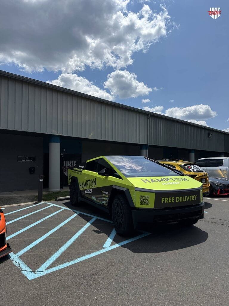 Tesla Cybertruck wrapped in green Hampton Market vinyl advertising, featuring 'Free Delivery' and QR codes, parked in a handicapped zone outside a commercial building with other custom cars in the background under a partly cloudy sky.